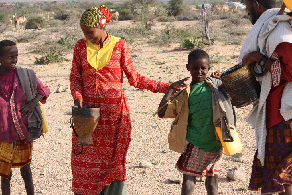 Two adults and two children in brightly patterned clothing carry wooden jugs across a sandy setting scattered with clusters of green grass.